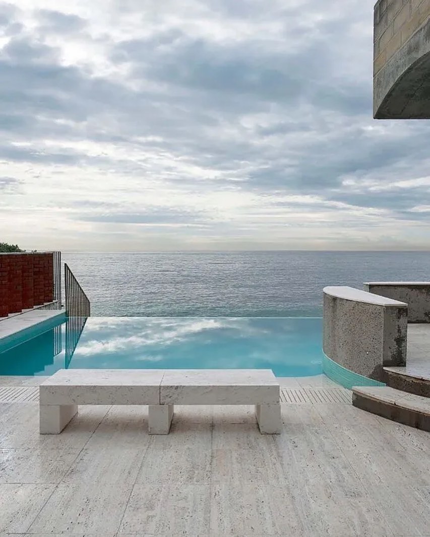 Infinity pool overlooking the ocean, featuring a stone bench on a travertine tile deck under a cloudy sky, blurring the lines between water and horizon.