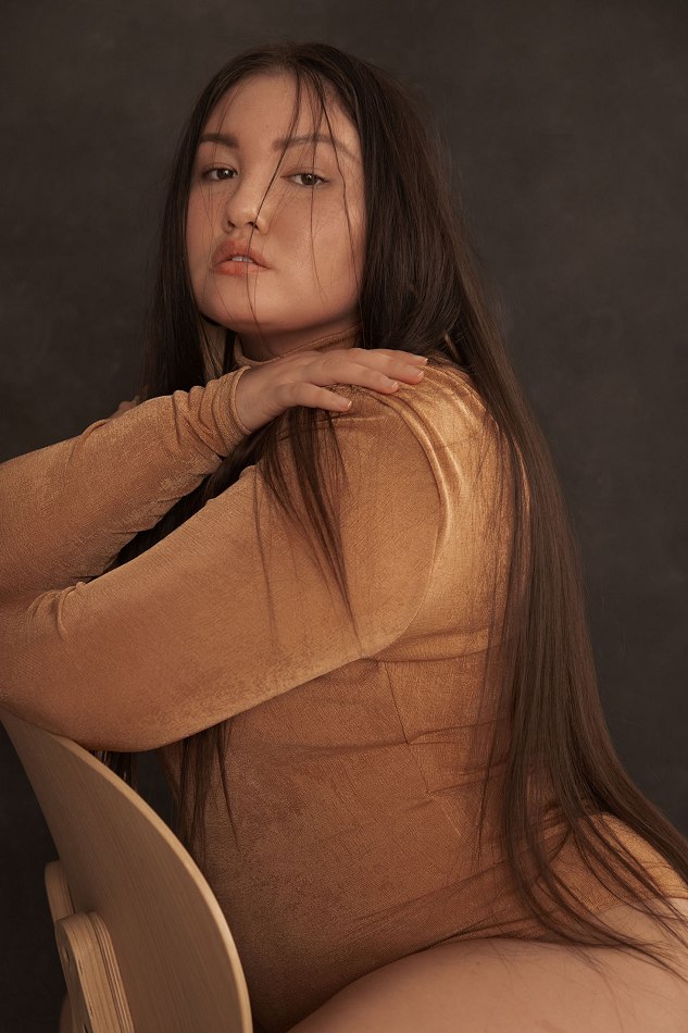 Portrait of an adult woman with long brown hair posing in a beige long-sleeve top on a chair against a dark grey background.