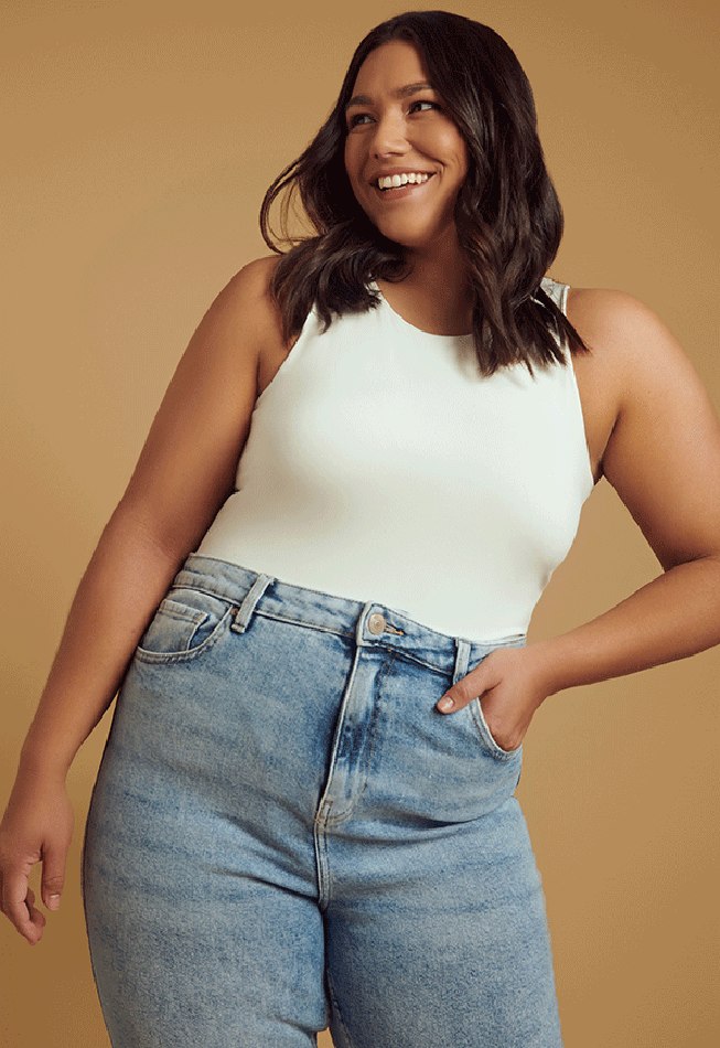 Portrait of a smiling woman wearing a light green tank top and jeans against a neutral background in a studio shot.