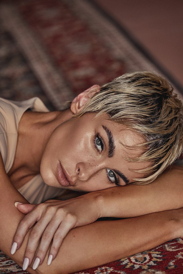 Close-up portrait of a young adult woman with short blonde hair and her arms crossed, laying on a patterned red and brown rug.