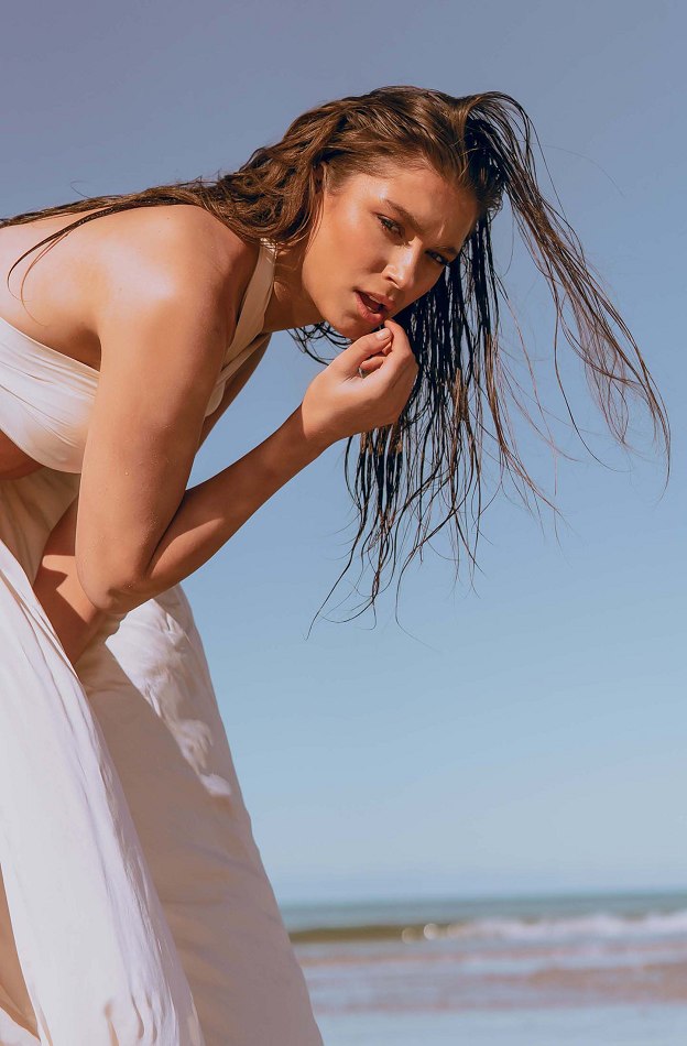 Woman in a swimsuit and pants poses on a sunny beach, with wet hair, against a blue sky and ocean backdrop.