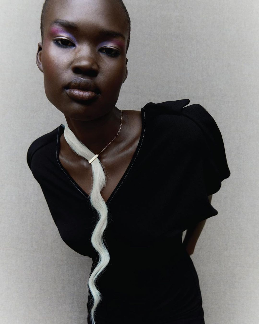 Close-up of an adult female model in a black dress with bright makeup and a decorative hairpiece posing indoors against a neutral wall.