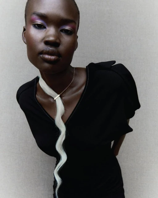 Close-up of an adult female model in a black dress with bright makeup and a decorative hairpiece posing indoors against a neutral wall.