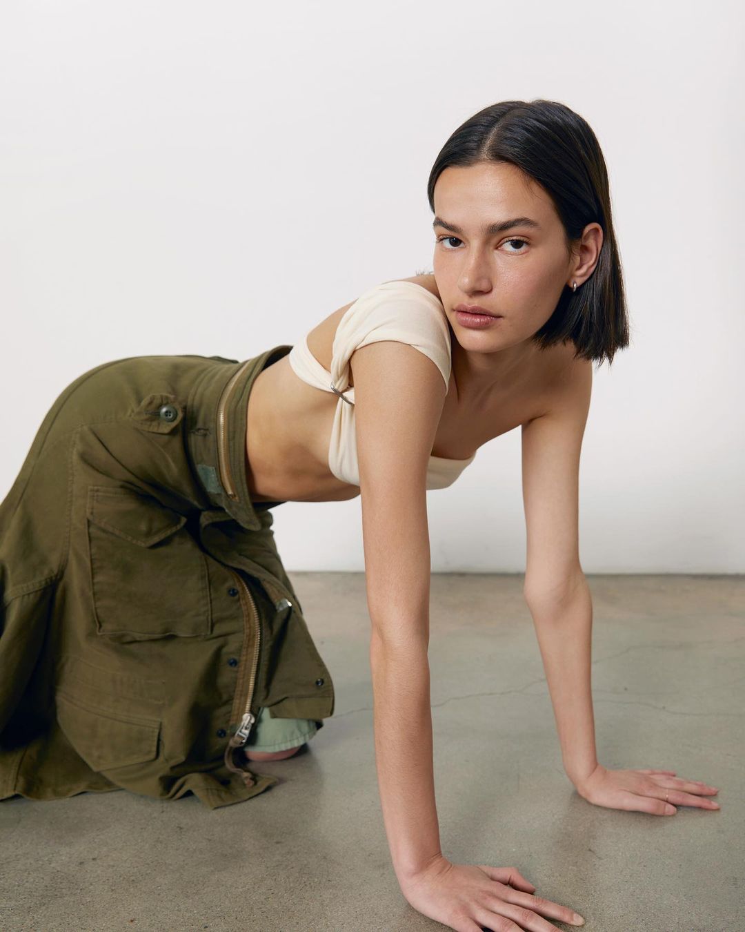 Brunette woman in a neutral-toned crop top and green cargo skirt poses kneeling on a concrete floor in a studio setting.