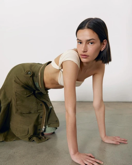 Brunette woman in a neutral-toned crop top and green cargo skirt poses kneeling on a concrete floor in a studio setting.