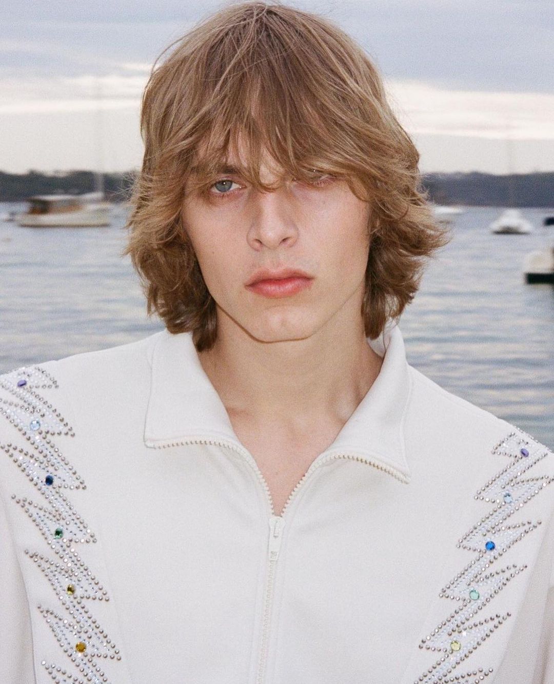 Close-up shot of a young adult with shoulder-length blond hair in a white embellished jacket against a water and sky background.