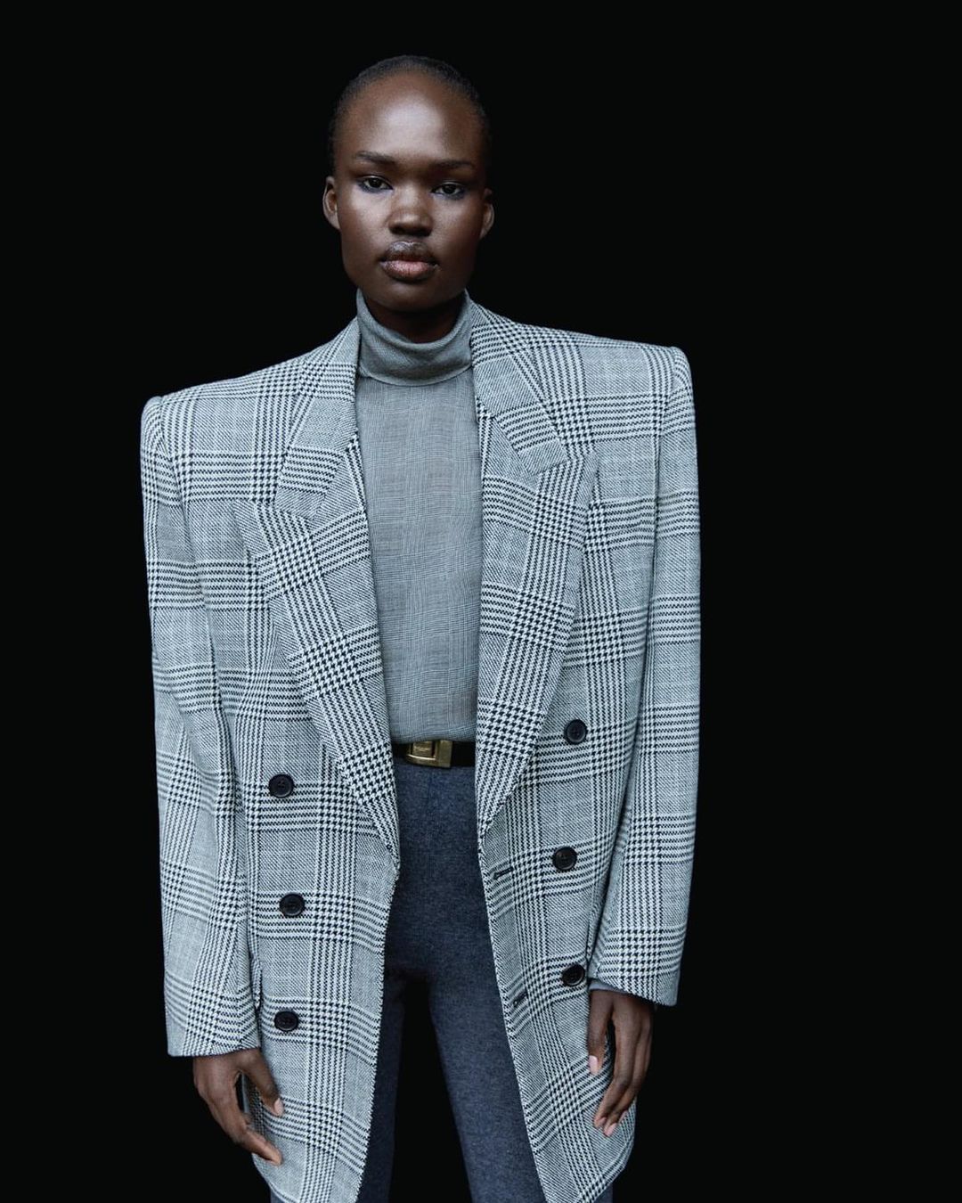 Woman in grey turtleneck and grey and white plaid blazer poses against a black background in a fashion shot.