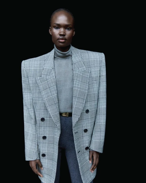 Woman in grey turtleneck and grey and white plaid blazer poses against a black background in a fashion shot.