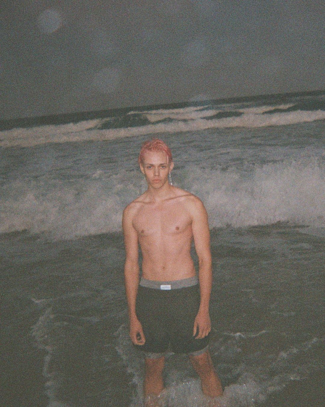 Shirtless person with pink hair stands in ocean surf with crashing waves and cloudy sky in background, medium shot, portrait orientation.