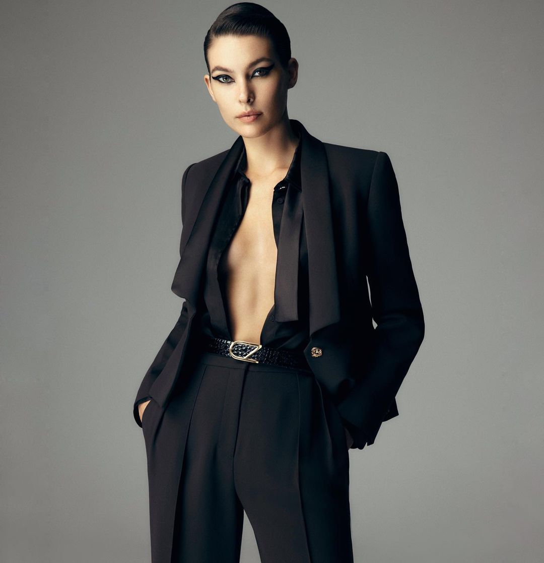 Fashion portrait of a stylish woman in a black blazer, top, and pants posing against a grey backdrop in a studio environment.