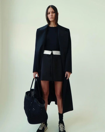 Model in dark long coat holding a black perforated tote bag against a pale background