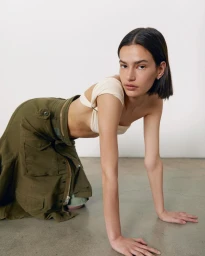 Model with dark bob hairstyle wearing an off-white top and olive green utility skirt kneeling on a concrete floor