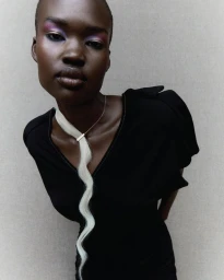 Close-up portrait of a model with short dark hair wearing a black dress and a long wavy blonde hair extension worn as a necklace.