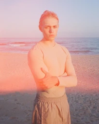 Blonde male model with arms crossed standing on a sandy beach by the ocean during sunset