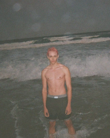Shirtless male model with pink hair standing in the surf of a dark ocean at night