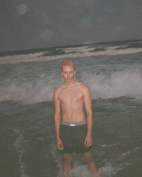 Shirtless male model with pink hair standing in the surf of a dark ocean at night