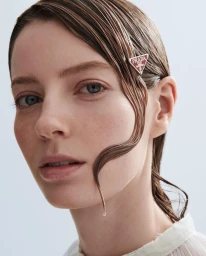 Close-up portrait of a woman with wet hair wearing a triangular Prada hair clip against a seamless white background.