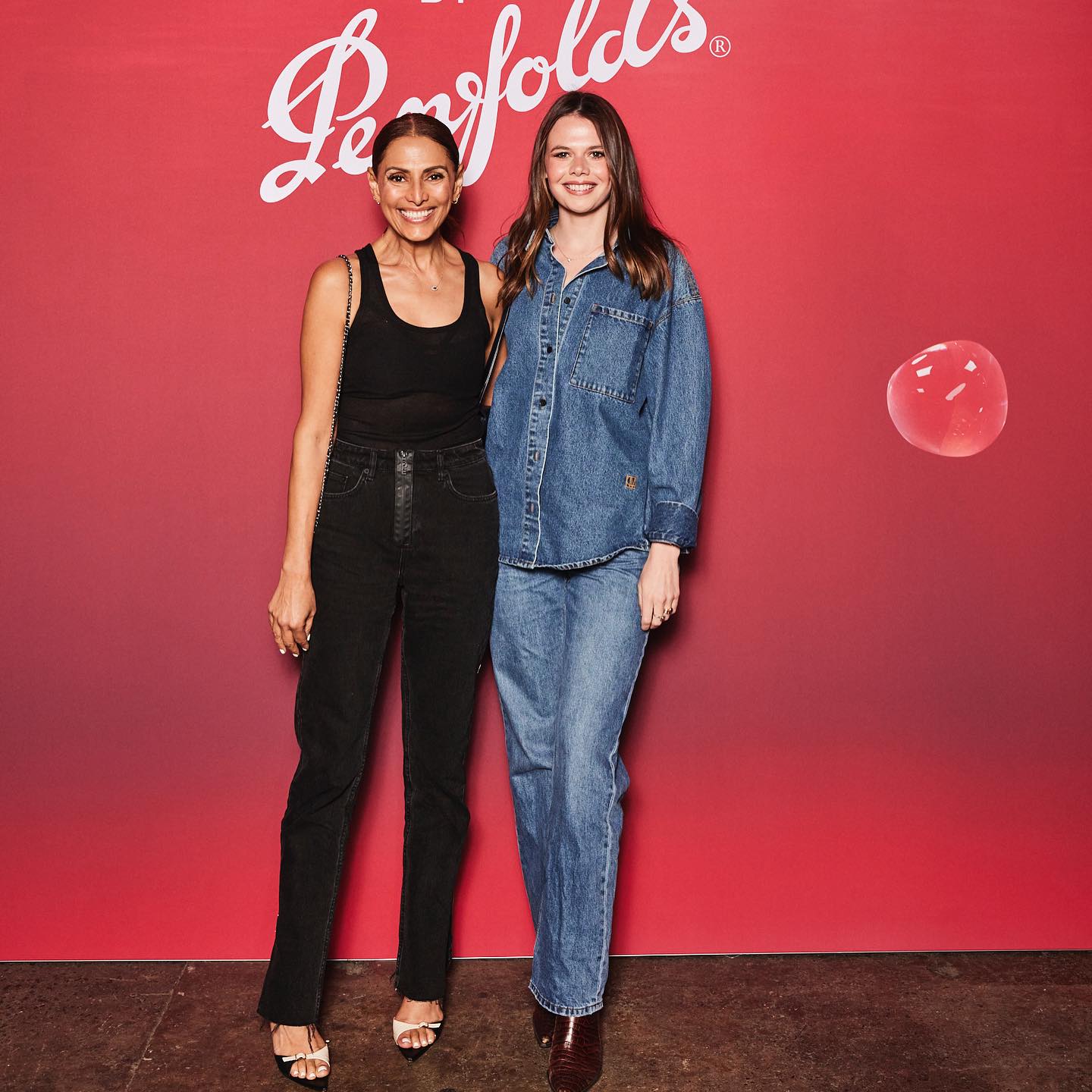 Two women pose in front of a Penfolds backdrop, one in denim, the other in black top and jeans, both smiling and fashionable.