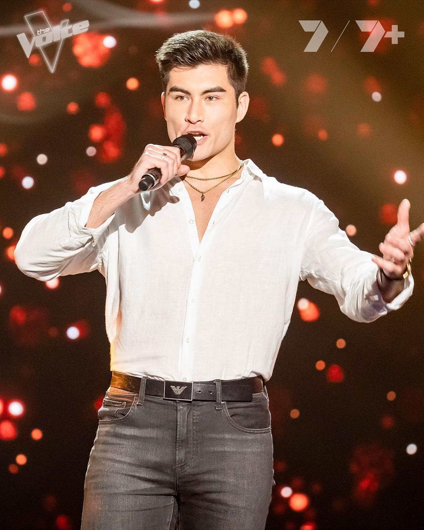 A man sings on stage with a microphone, wearing a white shirt and grey jeans against a blurred background with bokeh lights.