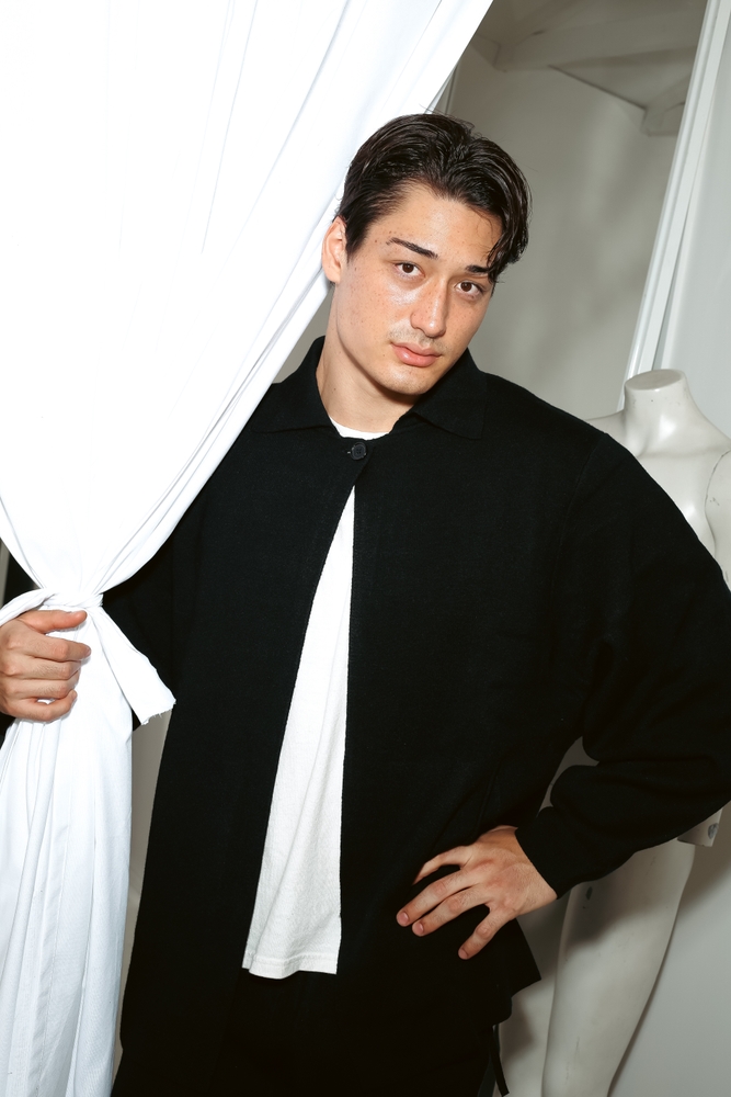 Portrait of a dark-haired adult male in a black jacket and white t-shirt, standing indoors with a white curtain and mannequin in the background.