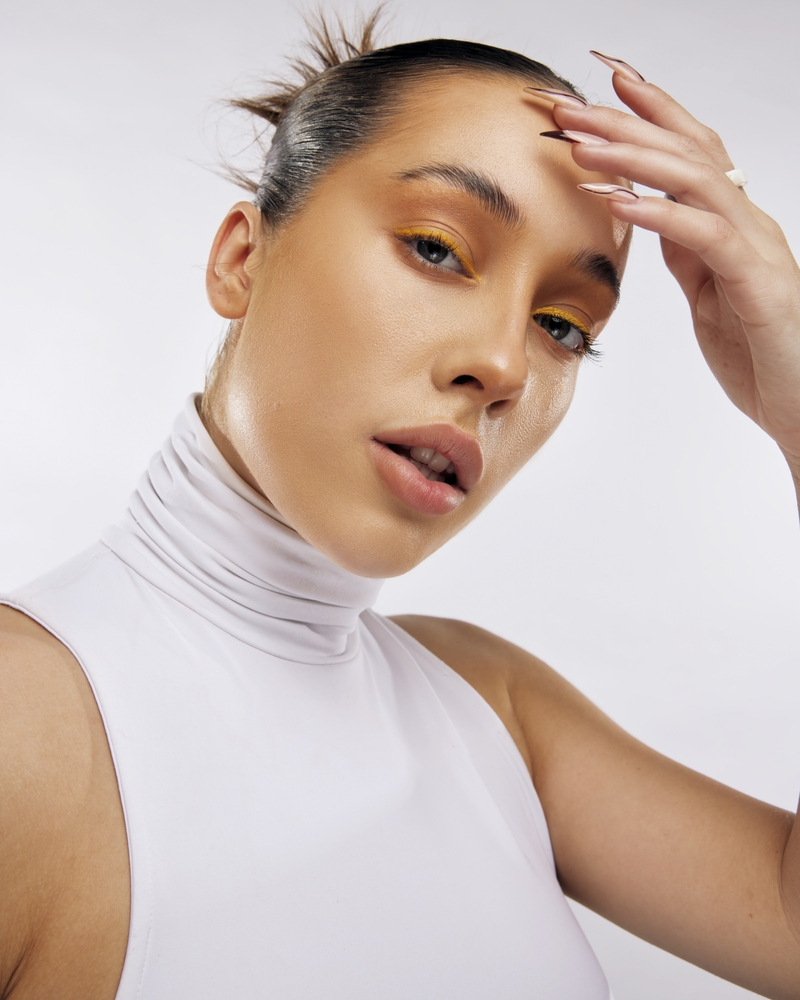 Close-up portrait of a young adult woman with light skin, styled hair, and yellow eyeshadow, posing indoors against a neutral backdrop.