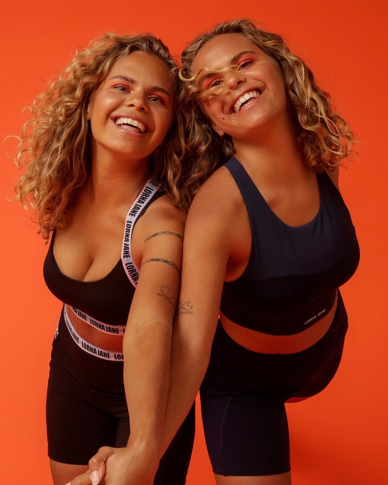 Two smiling women with curly hair pose in matching black and navy Lorna Jane sports bras and shorts against an orange background.
