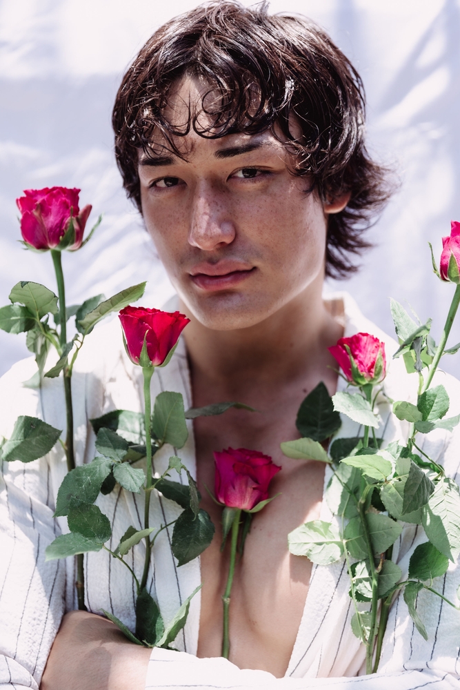 Close-up of an adult male model with dark hair, posing amid red roses against a white background in a fashion or beauty context.