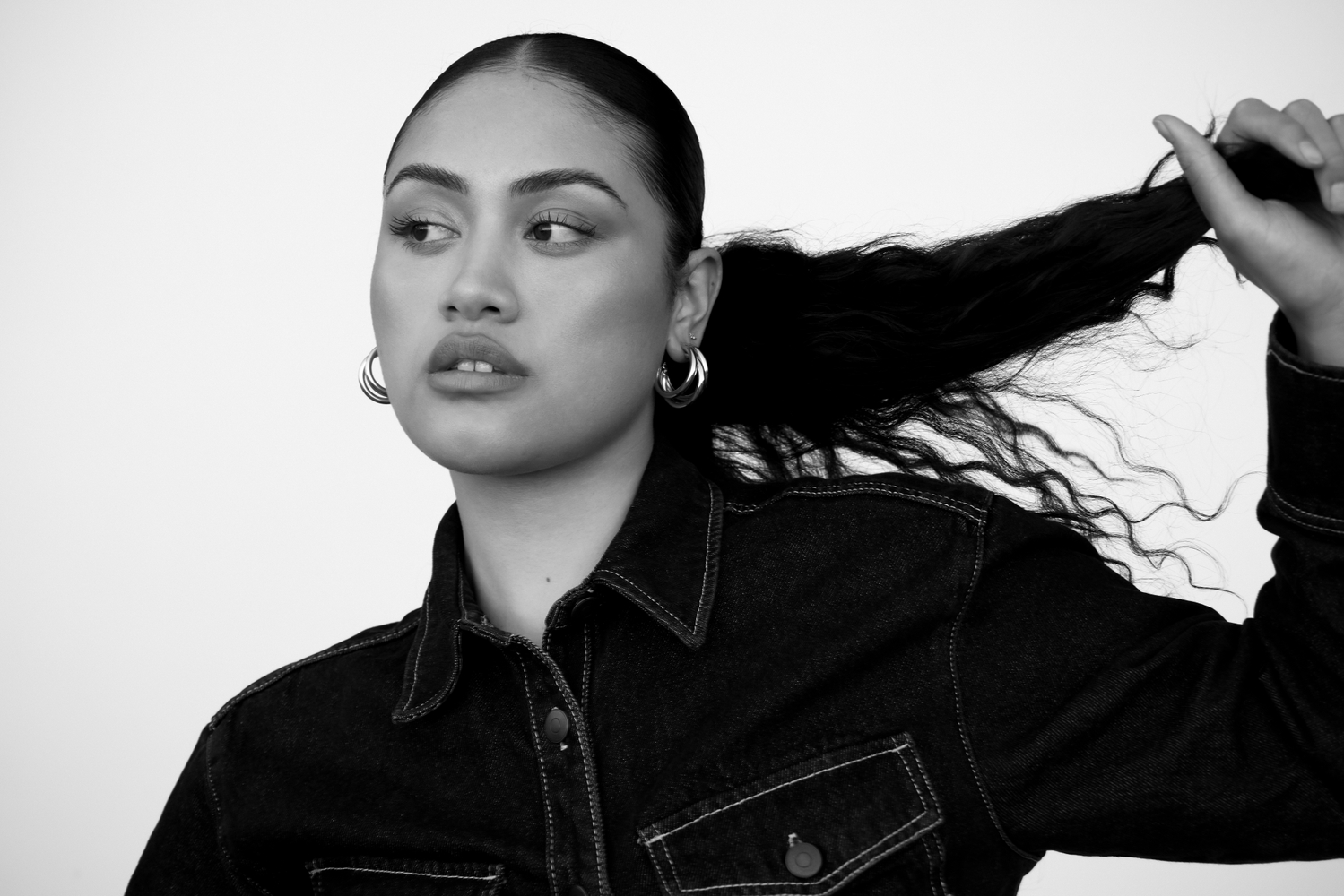 Portrait of a woman with dark hair pulled back, posing in front of a white background, wearing denim shirt and hoop earrings.