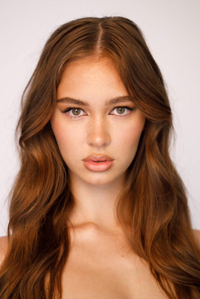 Close-up portrait of a beautiful woman with long auburn wavy hair, fair skin, light eyes, and subtle makeup against a white background.