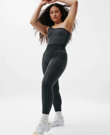 Model in dark grey one-shoulder bodysuit and white sneakers posing against a white background