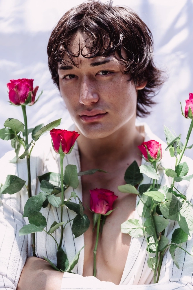 Young man with dark wet hair holding red roses wearing a striped white shirt against a white backdrop.