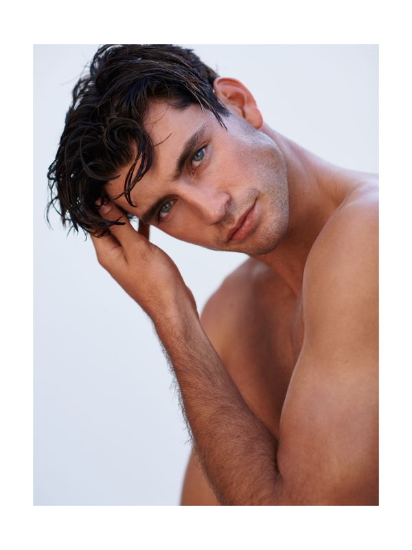 Close-up studio portrait of a shirtless adult male with dark hair, light eyes, and tanned skin, looking at the camera, hand on head.