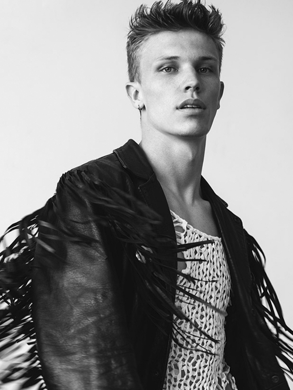 Portrait of a young adult male model in a fringed jacket and patterned shirt against a plain background in a studio shot.