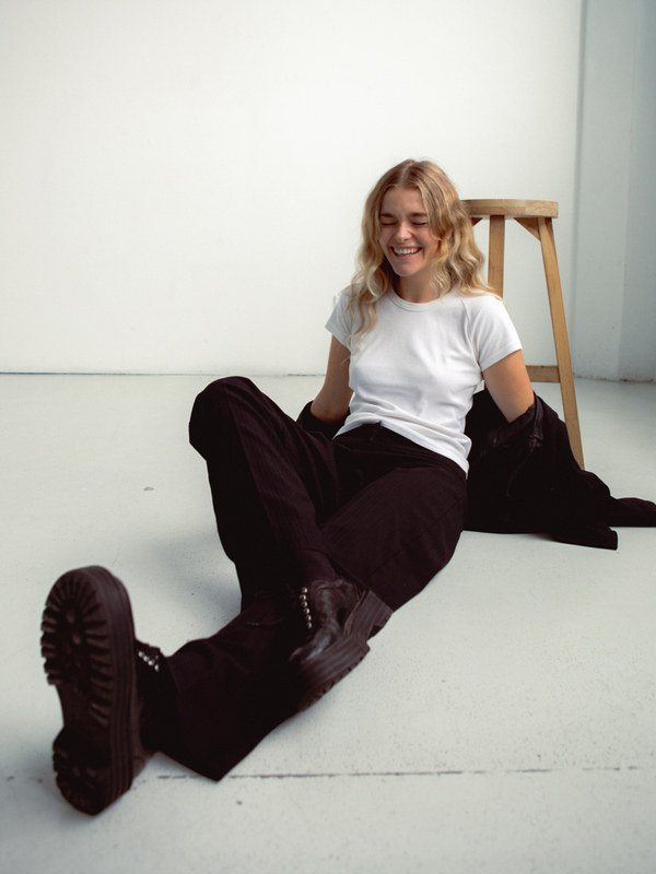 A woman with long blonde hair smiles while sitting on the floor in a white studio with black shoes, pants, and a white t-shirt.