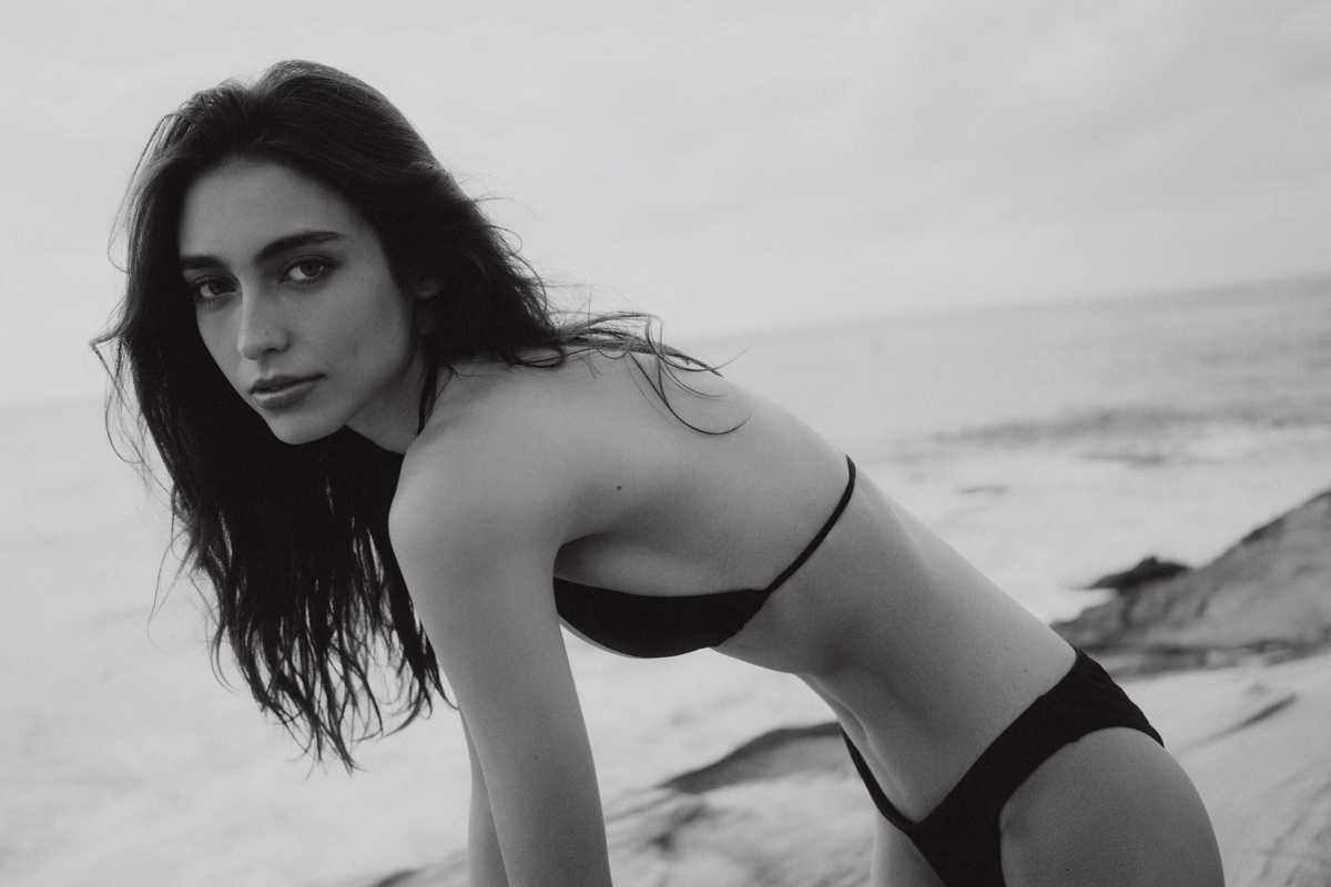 Monochrome portrait of a woman in a bikini leaning forward on a beach with a neutral expression, captured in a medium shot.