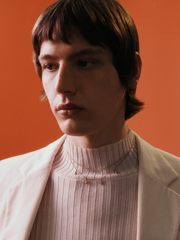 Portrait of a young adult male with dark hair, wearing a turtleneck and blazer, accessorized with delicate necklaces against an orange backdrop.