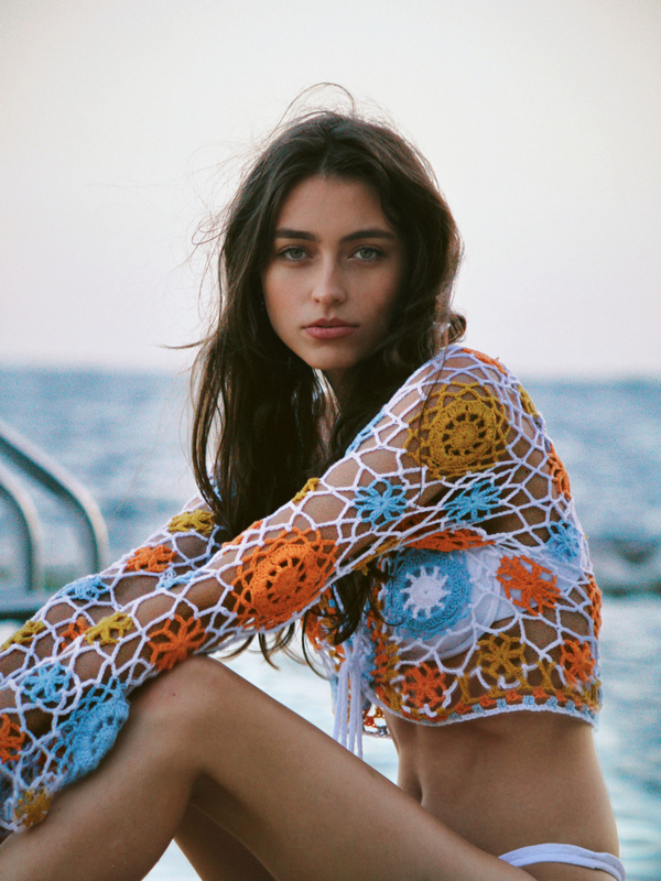 A woman with long, dark hair is posed near a pool. She's wearing a crocheted crop top with a view of the water in the background.