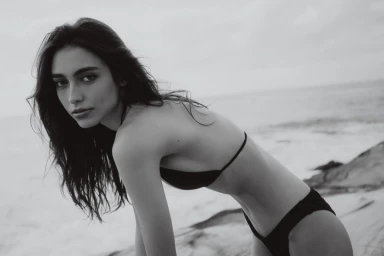 Model in a black bikini posing on a sandy beach under an overcast sky, captured in monochrome.