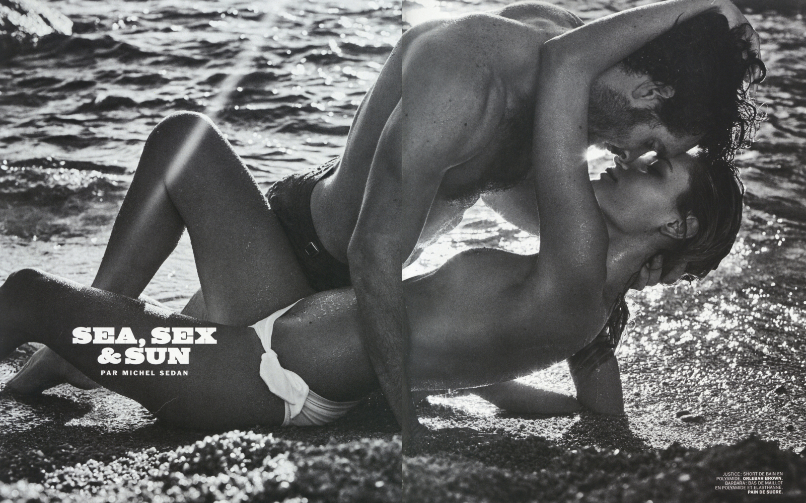 Black and white image of a shirtless couple embracing on a sandy beach, with ocean water in the background, under bright sunlight.