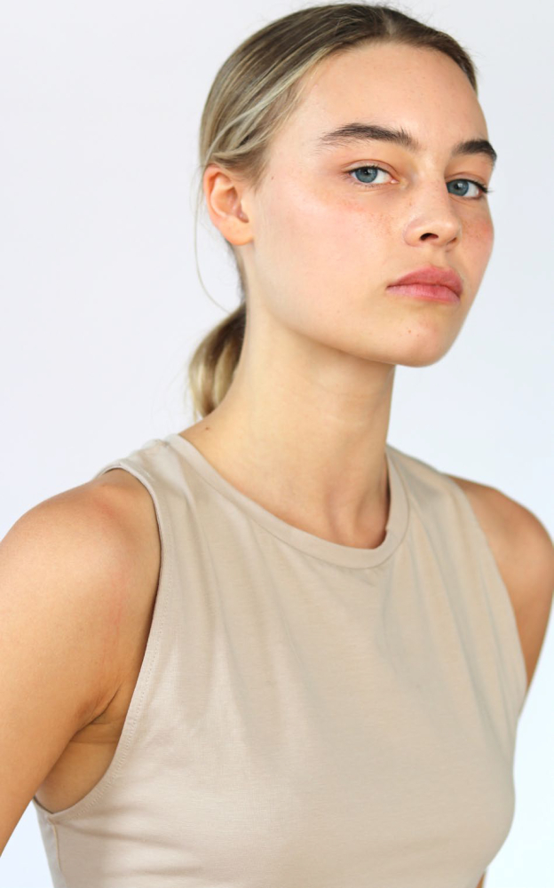 Portrait of a young adult woman with blonde hair pulled back, wearing a tan tank top, posing against a white background in a photo studio.