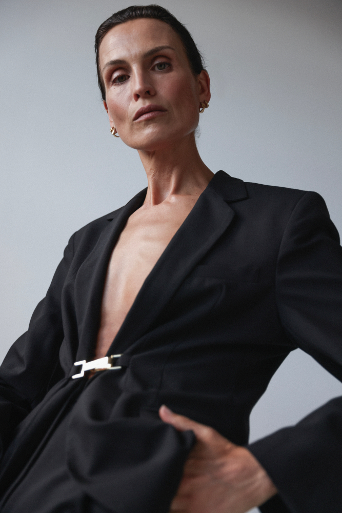 Elegant woman in a black blazer with a gold clasp posing against a grey background in a fashion photography style portrait.