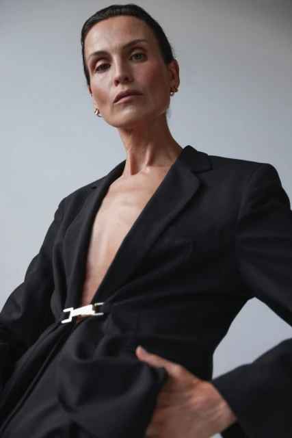Elegant woman in a black blazer with a gold clasp posing against a grey background in a fashion photography style portrait.