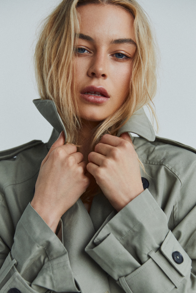 Close-up portrait of a blonde adult female in a khaki trench coat against a neutral background in a fashion photography style.