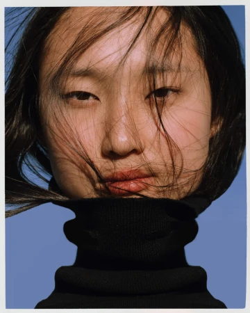 Close-up portrait of an East Asian woman with dark hair blowing across her face wearing a black turtleneck against a clear blue sky