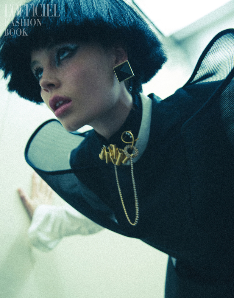 Eye-level fashion shot of a young woman with a blunt bob haircut wearing a black top and bold square earrings, posing near a wall.
