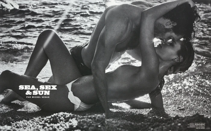Black and white editorial photograph of a couple embracing closely on a sandy beach near water with sunlight reflecting off the surface.