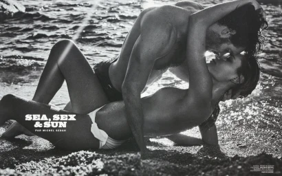 Black and white editorial photograph of a couple embracing closely on a sandy beach near water with sunlight reflecting off the surface.