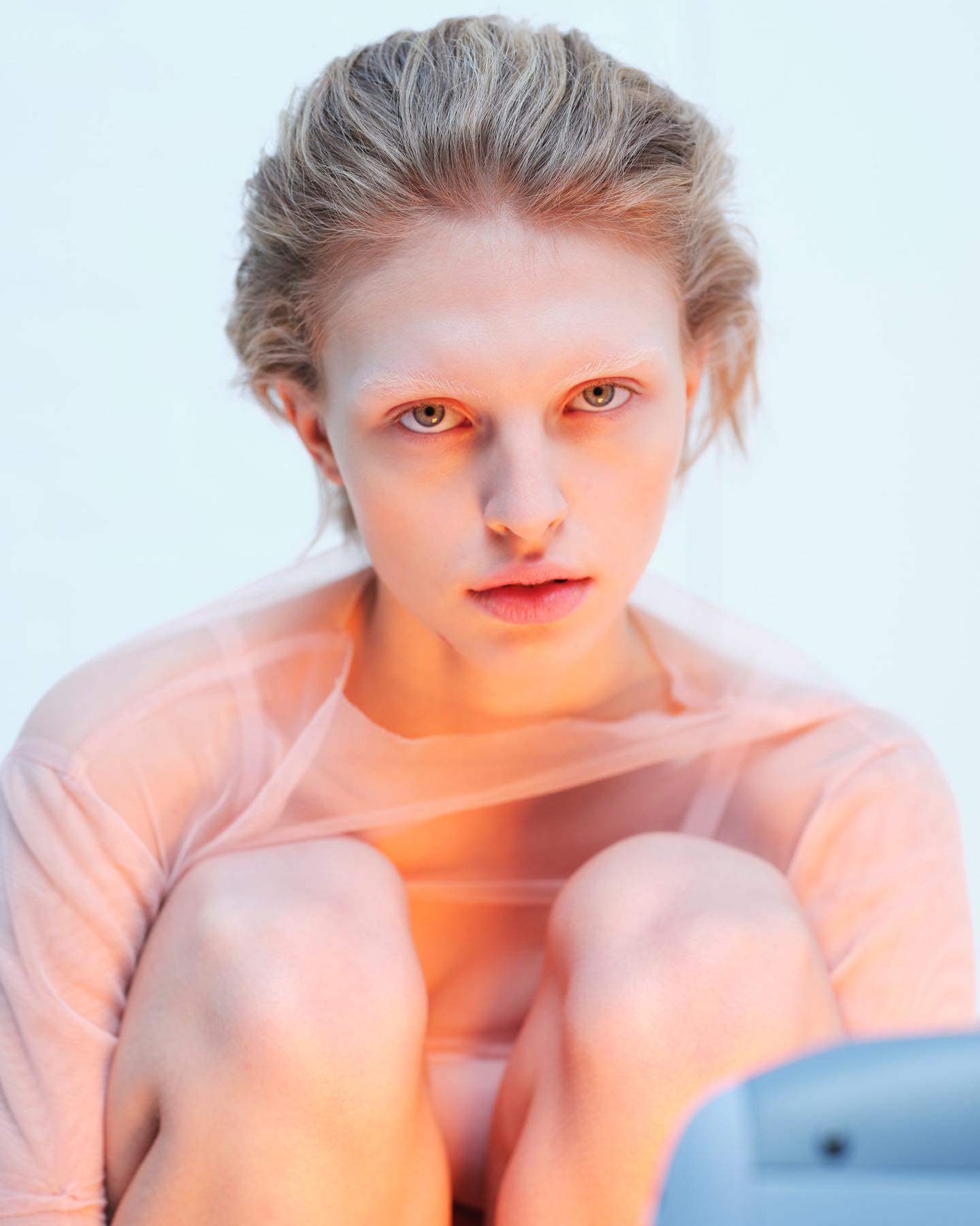 Close-up of a young adult woman with pale skin and light blonde hair, wearing a sheer peach top, and looking directly at the camera.