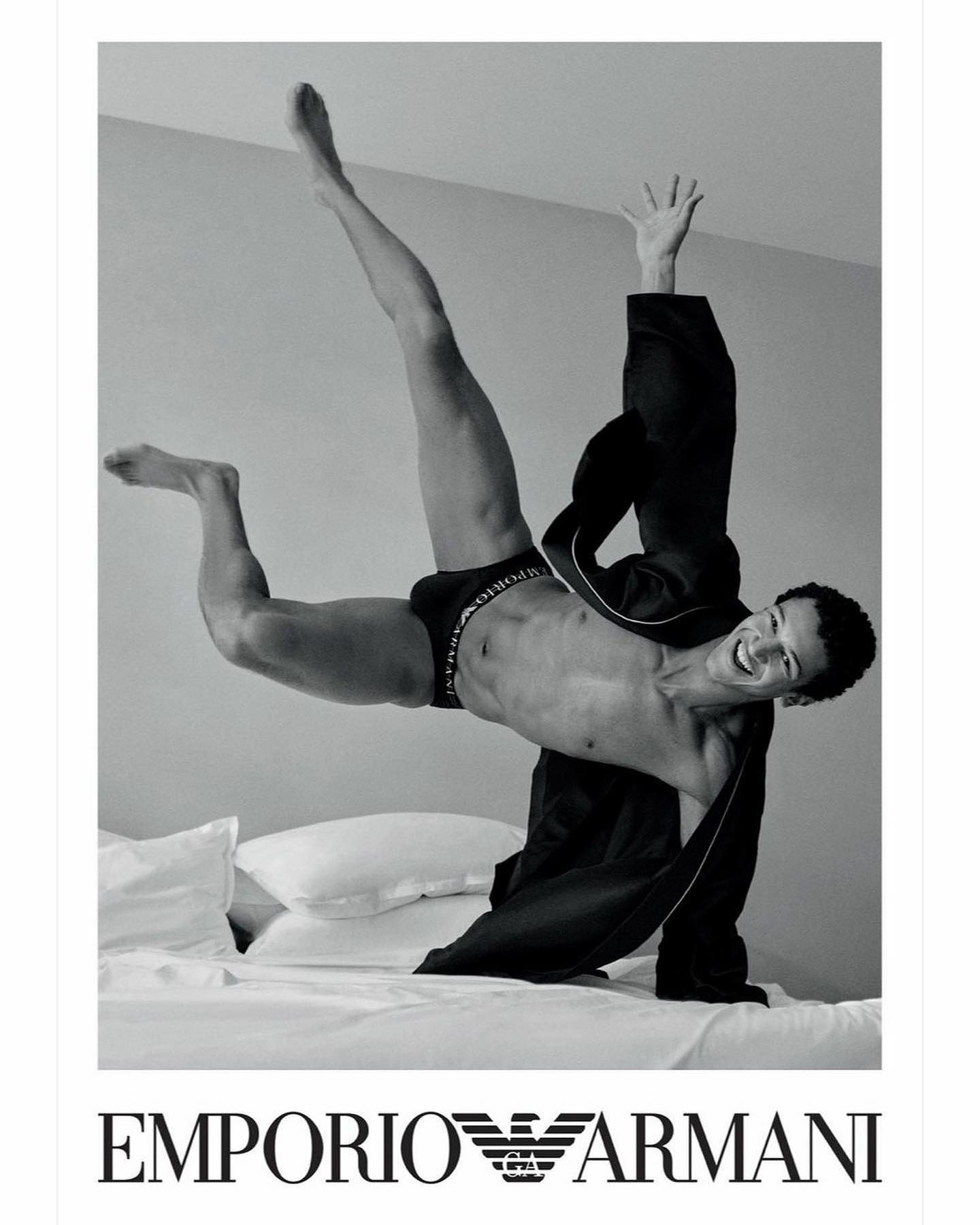 A male fashion model strikes a dynamic acrobatic pose on a bed in an Emporio Armani underwear advertisement shot in black and white.