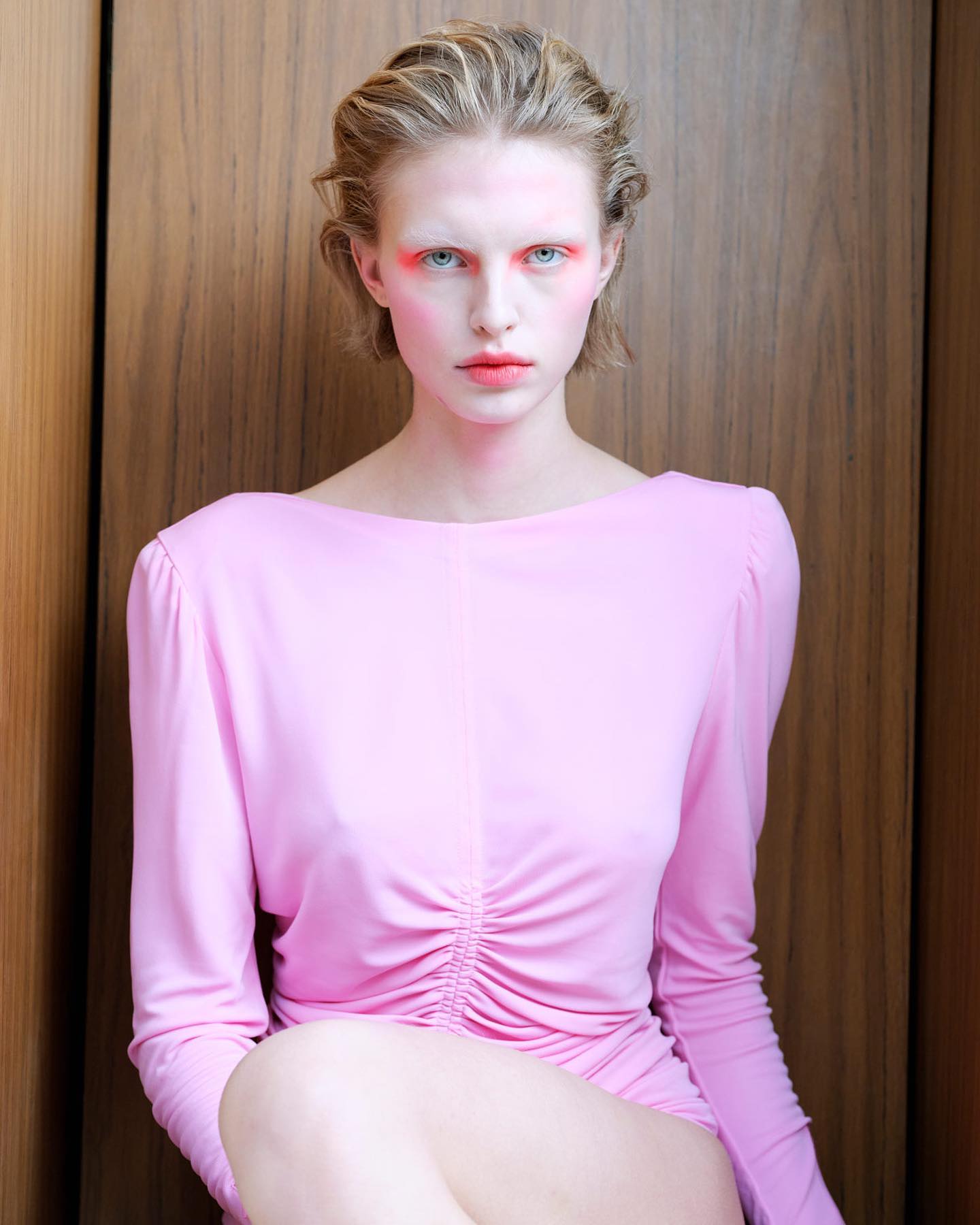 Woman with blonde hair and red makeup poses in a pink long-sleeve dress against a wood background in a fashion or beauty setting.